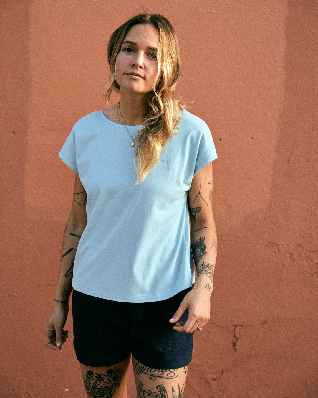 A person with long hair stands in front of a textured peach-colored wall, wearing a light blue t-shirt and Liesl - Womens Chino Shorts - Blue by Saltrock made from 100% cotton, with visible tattoos on both arms.