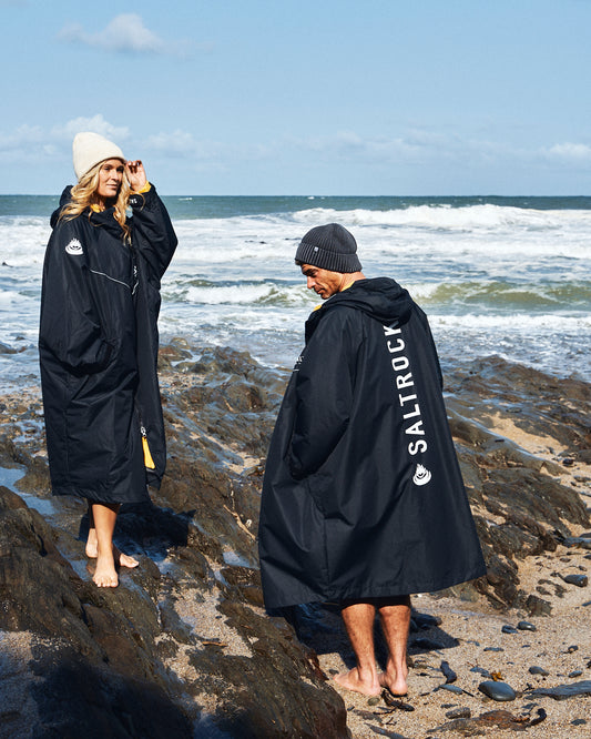 Two people in Saltrock's 3 in 1 Recycled Changing Robe in Black/Yellow, crafted from recycled material, stand barefoot on a rocky beach with waves behind them, wearing beanies as they enjoy their versatile robes.