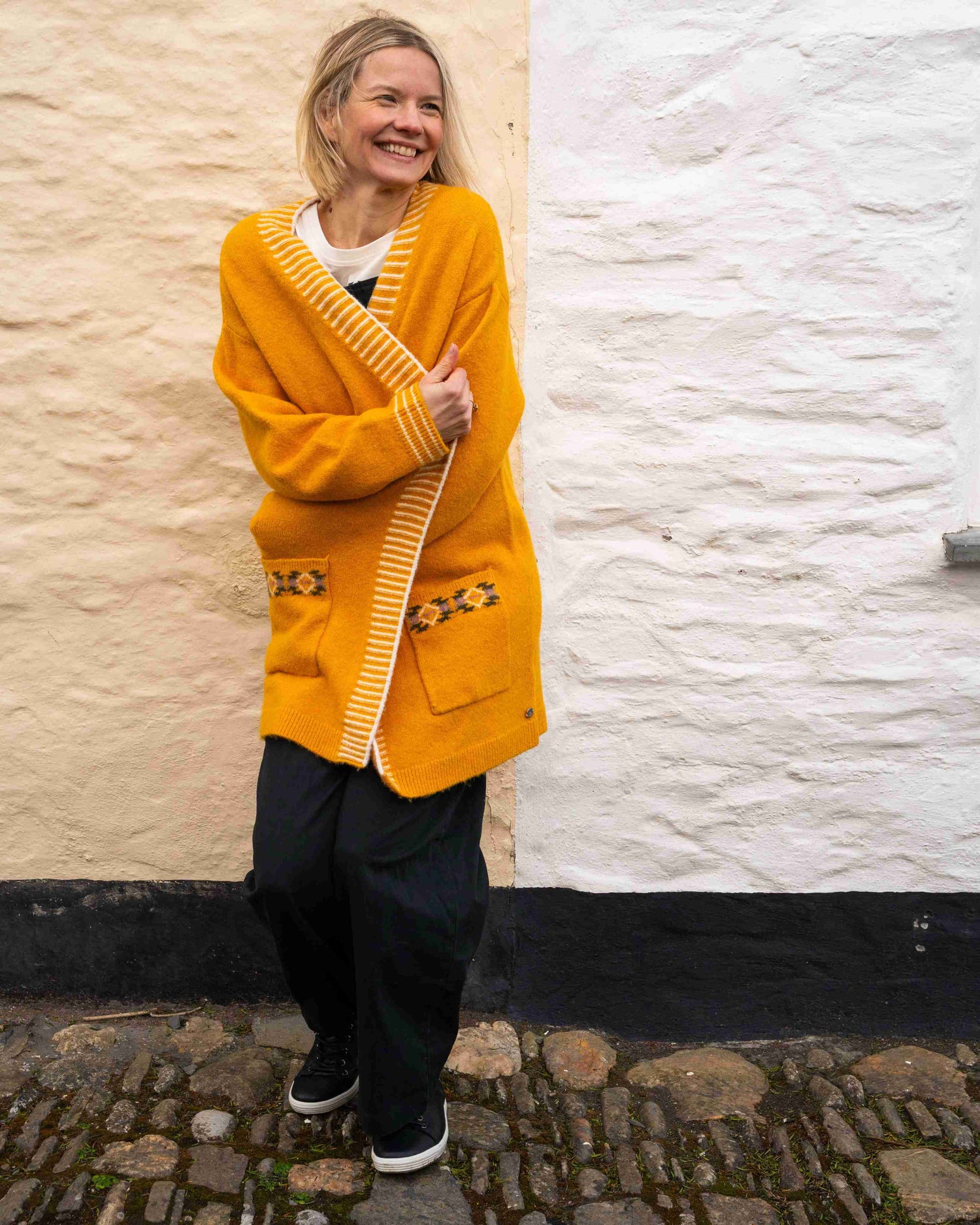 Wearing the Saltrock Coraline Longline Knitted Cardigan in Yellow, a woman smiles on a cobblestone path against a textured white and beige wall, embracing its open-front style with pockets and striped trim for added flair.