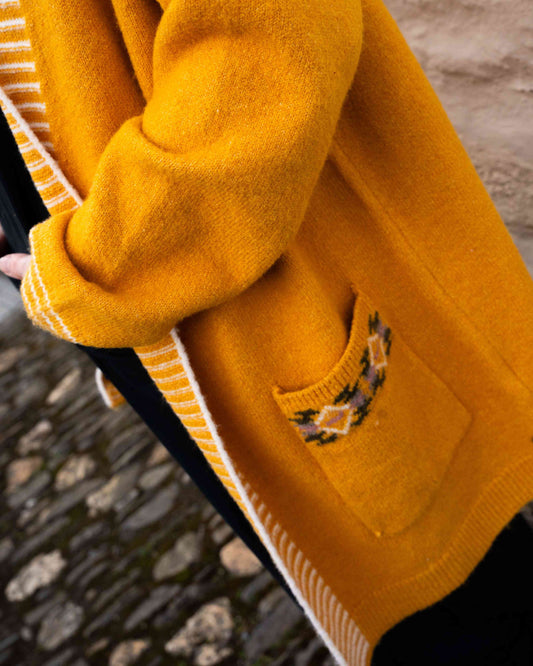 Someone dons the Saltrock Coraline, a mustard yellow longline knitted cardigan, featuring a floral patterned pocket and an open-front style, as they stand on a cobblestone path.