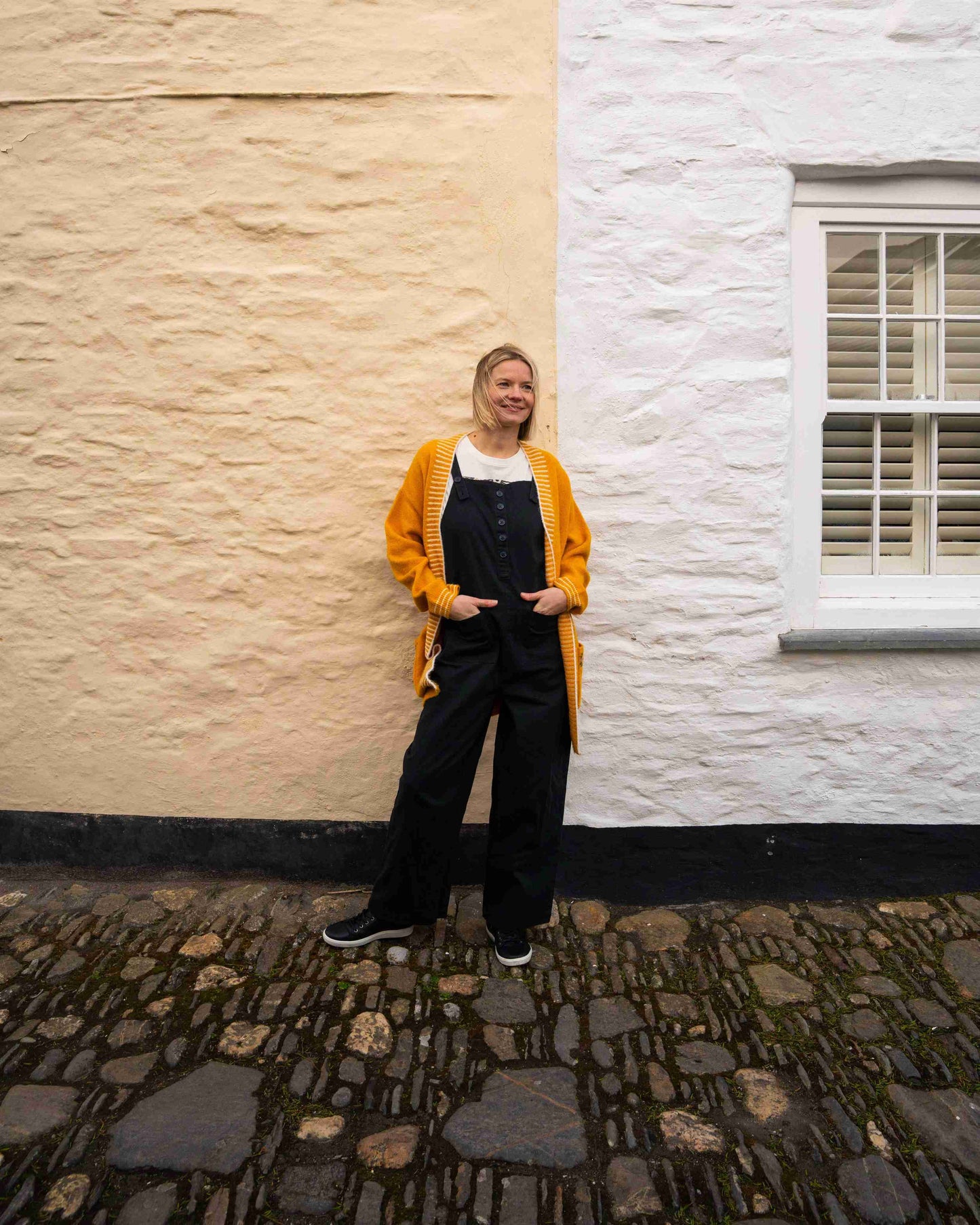 A person in a Saltrock Coraline yellow longline knitted cardigan and black overalls stands by a two-toned stone wall near a window with white shutters, with the cobblestone ground adding charm to the scene.