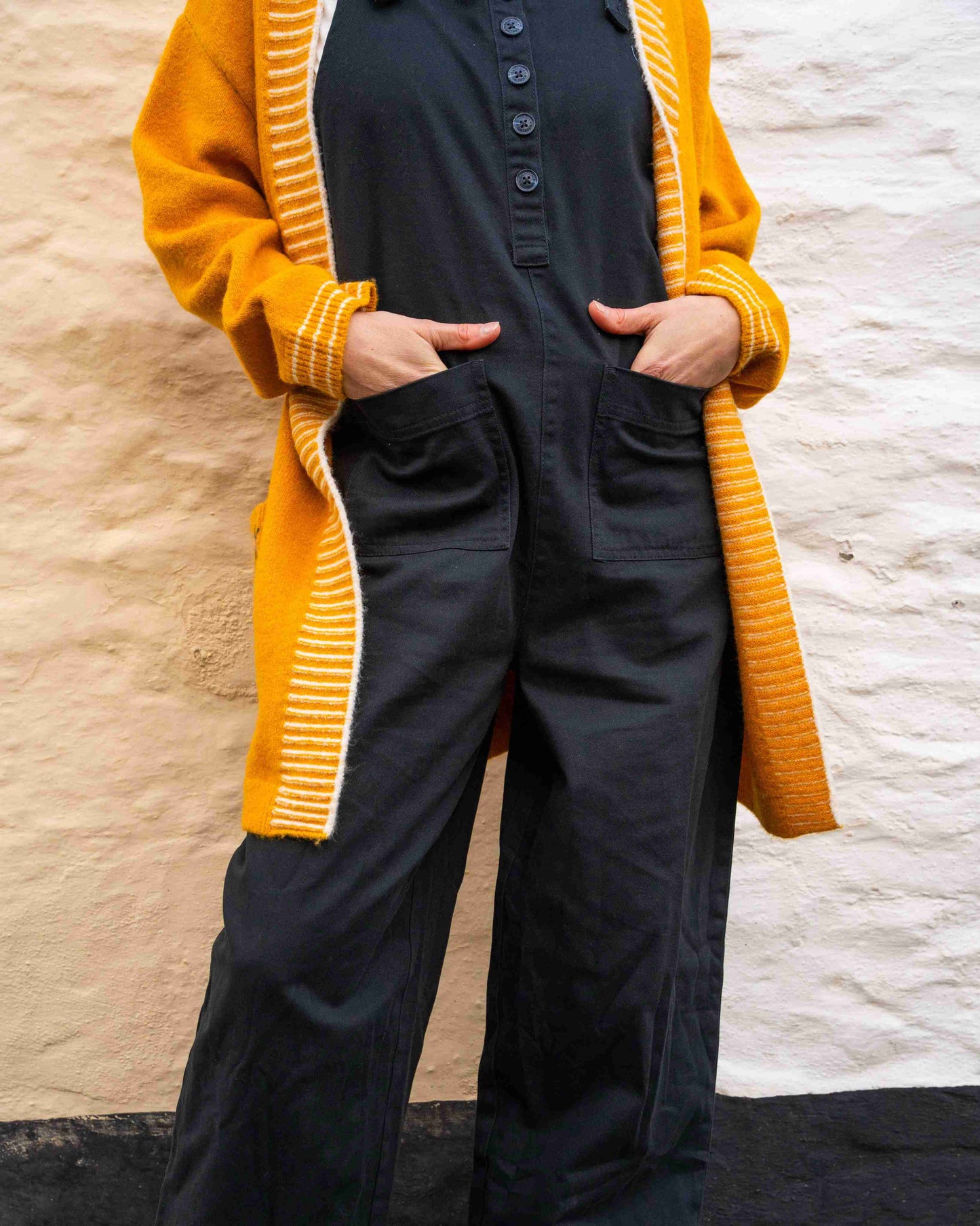 A person in the Saltrock Coraline women's longline knitted cardigan in yellow and a black jumpsuit stands with hands in pockets against a two-tone wall.