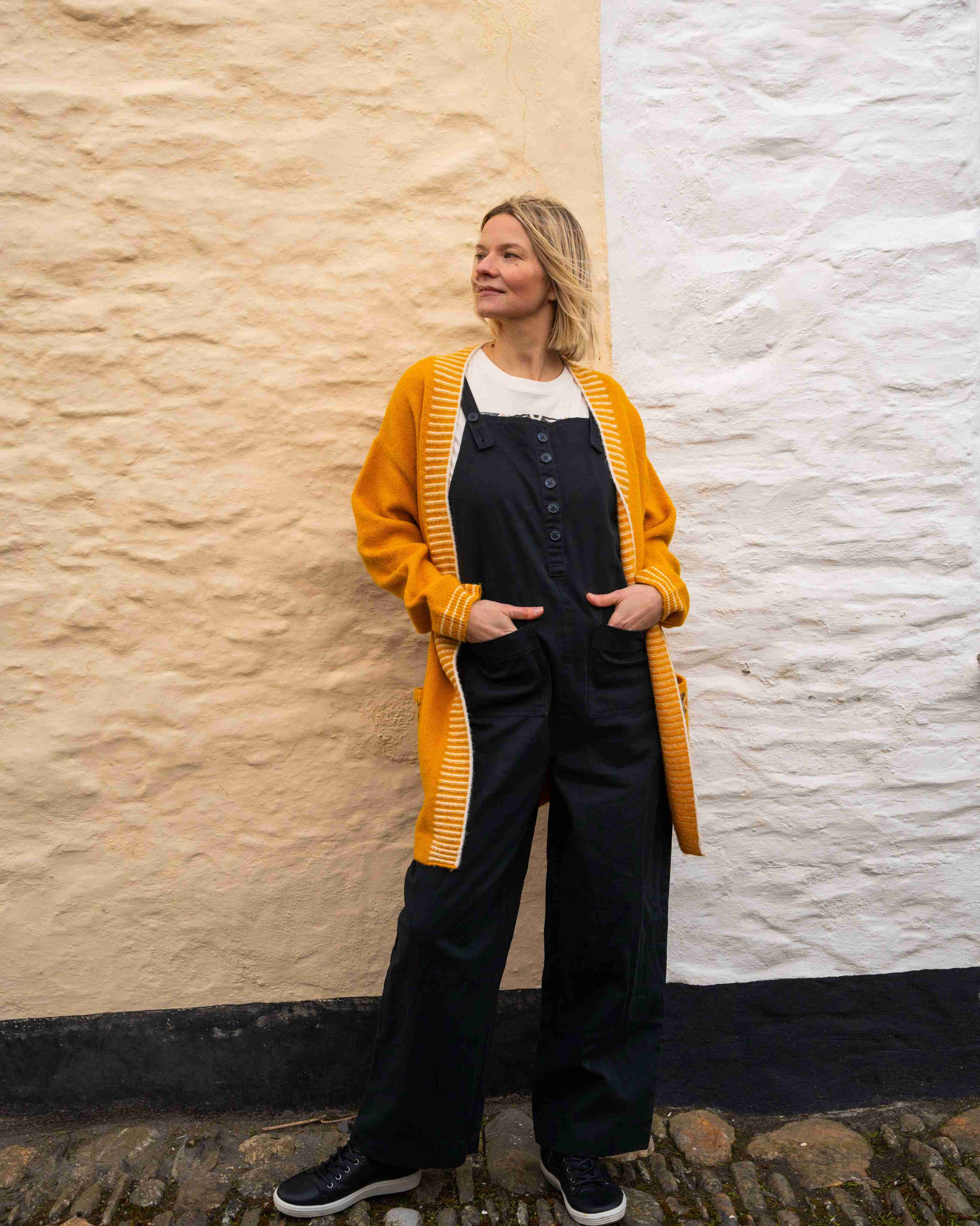 A person stands before a textured wall, wearing the Saltrock Coraline yellow longline knitted cardigan over a black jumpsuit with hands in pockets.