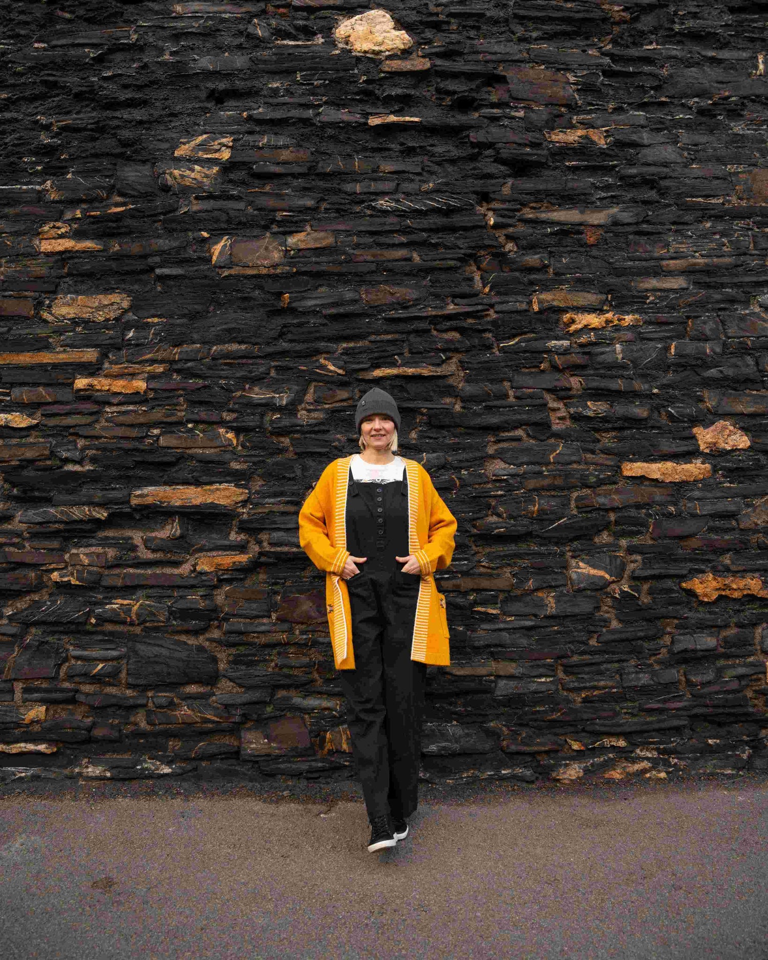 A person in a yellow Coraline longline knitted cardigan by Saltrock, paired with a black outfit, poses against a textured stone wall, hands in pockets, wearing a hat and sneakers.