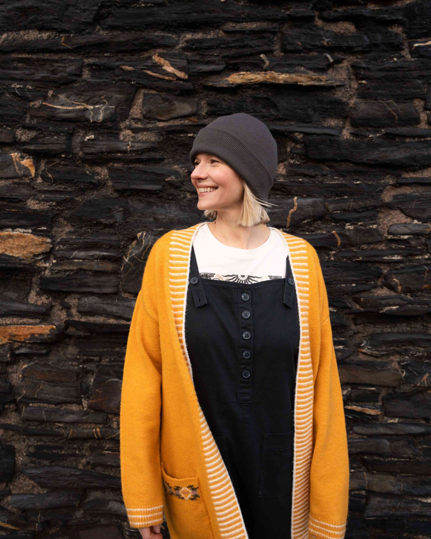 A person wearing a Saltrock Coraline Women's Longline Knitted Cardigan in yellow and a gray hat smiles in front of a brick wall.