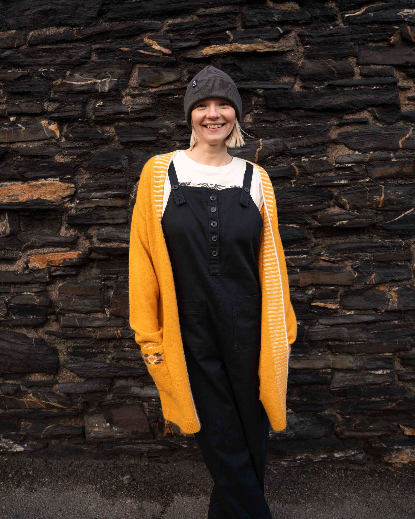 A person wearing the Saltrock Coraline Women's Longline Knitted Cardigan in yellow, black overalls, and a gray beanie stands smiling in front of a stone wall.