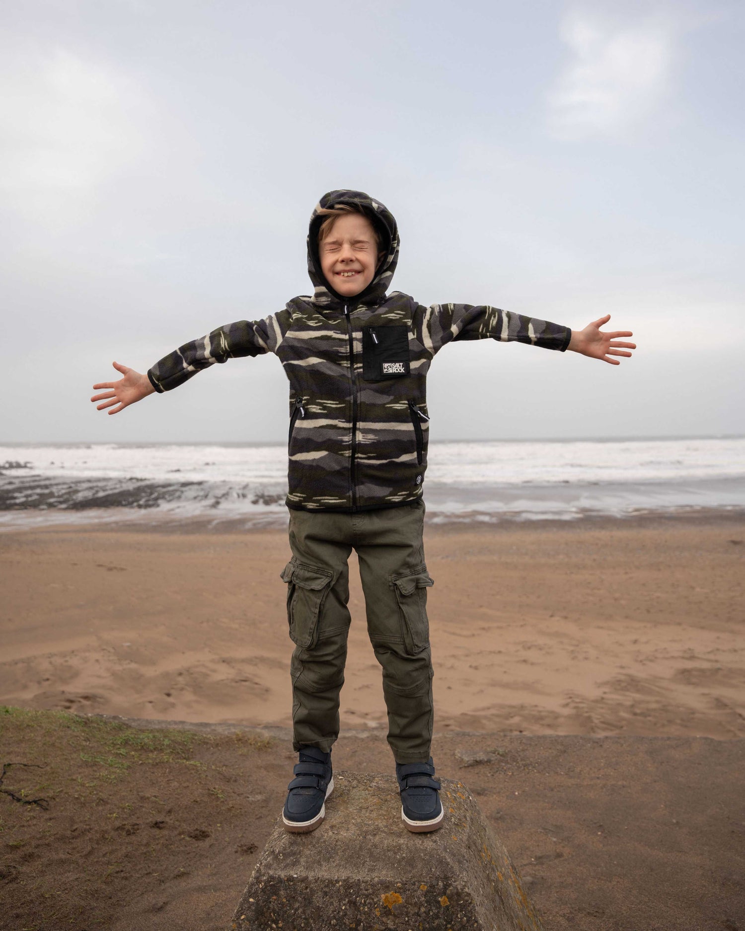 A child in a Saltrock Camo Stripe Kids Recycled Micro Fleece jacket stands on a concrete block at the beach, arms spread wide, enjoying the ocean breeze and cloudy sky.