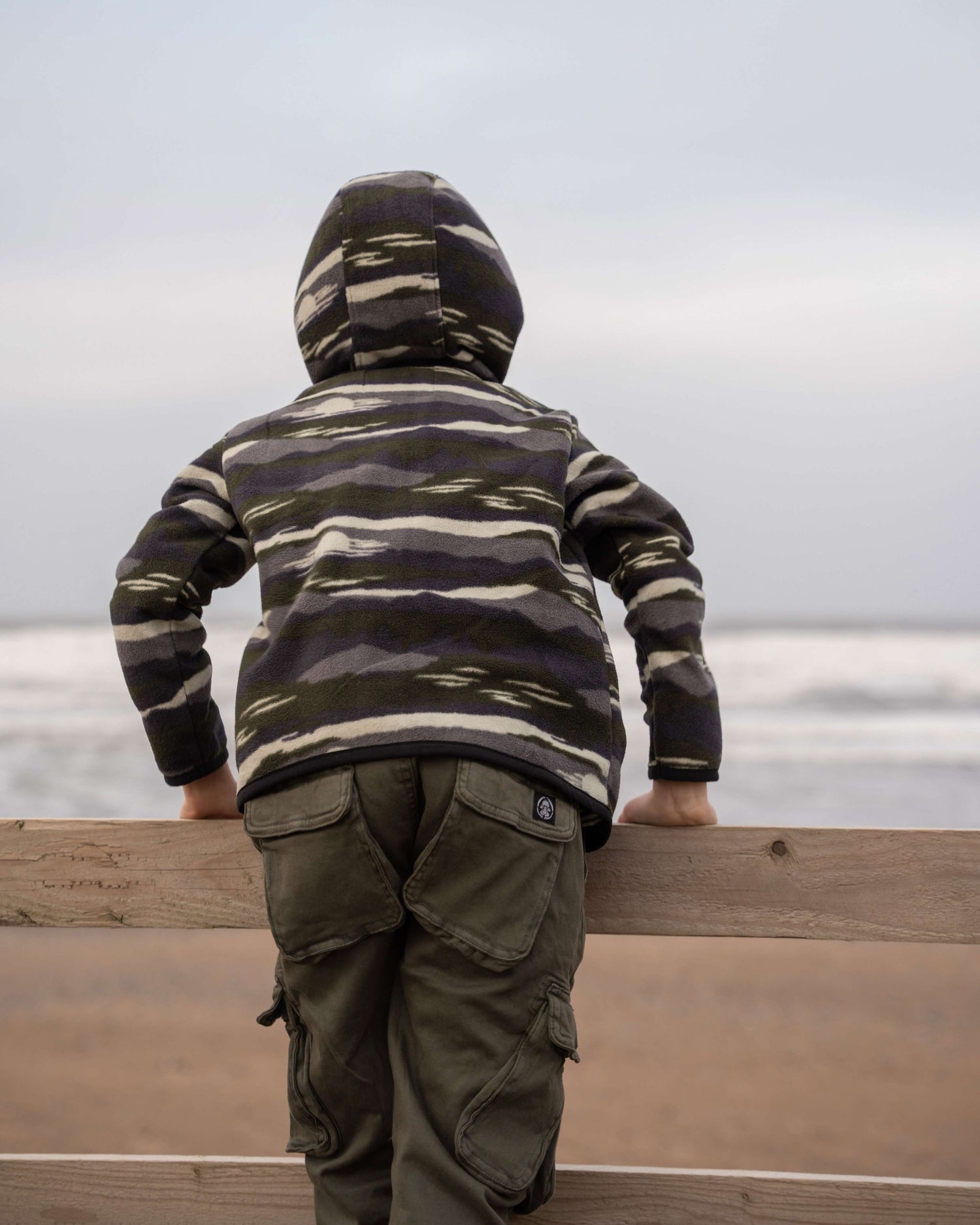 A child in a Saltrock Camo Stripe Kids Recycled Micro Fleece hoodie and green pants leans on a wooden railing, gazing at the ocean under a cloudy sky.