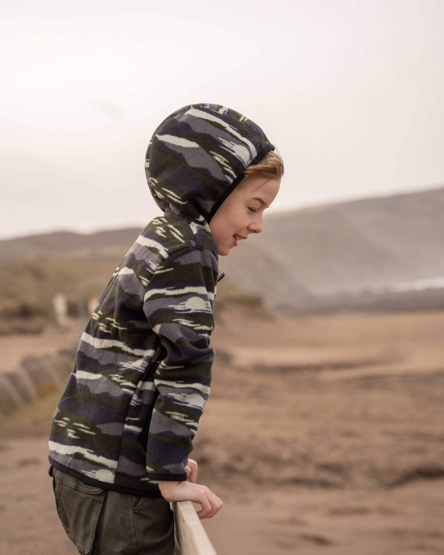 A child in a Saltrock Camo Stripe Kids Recycled Micro Fleece Hoodie with a chest pocket leans over a railing, gazing at the sandy beach and hills beyond.