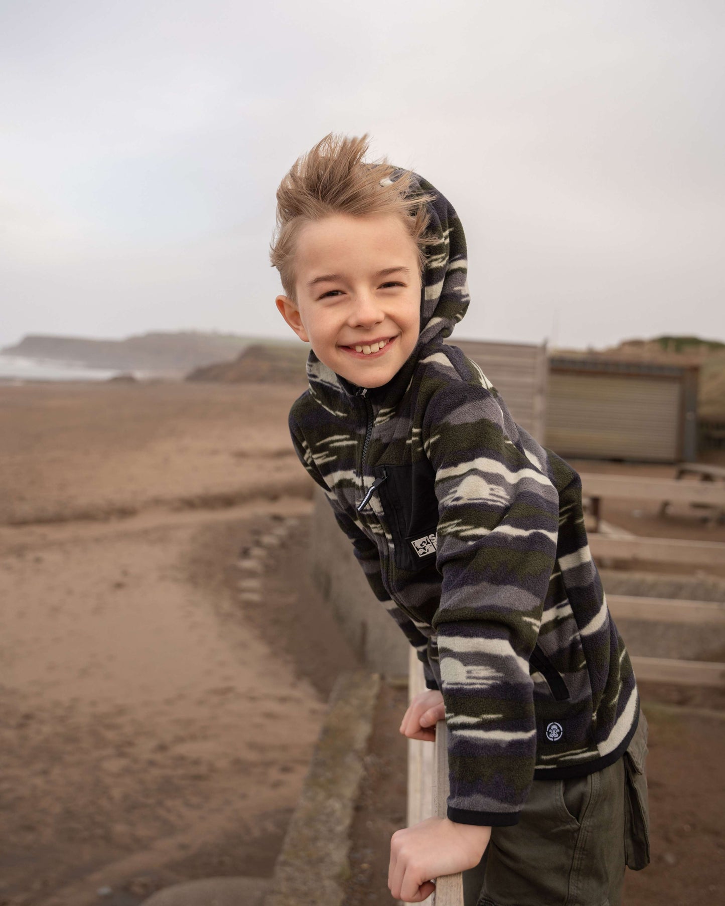 A child in Saltrock's Camo Stripe - Kids Recycled Micro Fleece, featuring a chest pocket, smiles while leaning on a beach railing. The sea and cloudy sky provide a serene backdrop for this eco-friendly green hoodie made from recycled materials.