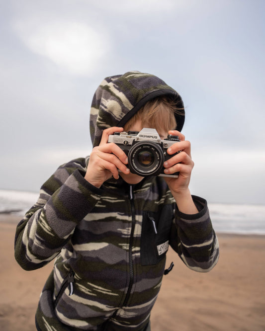 A child wears Saltrock's Camo Stripe - Kids Recycled Micro Fleece in Green, crafted from recycled polyester, while holding a camera on an overcast beach.