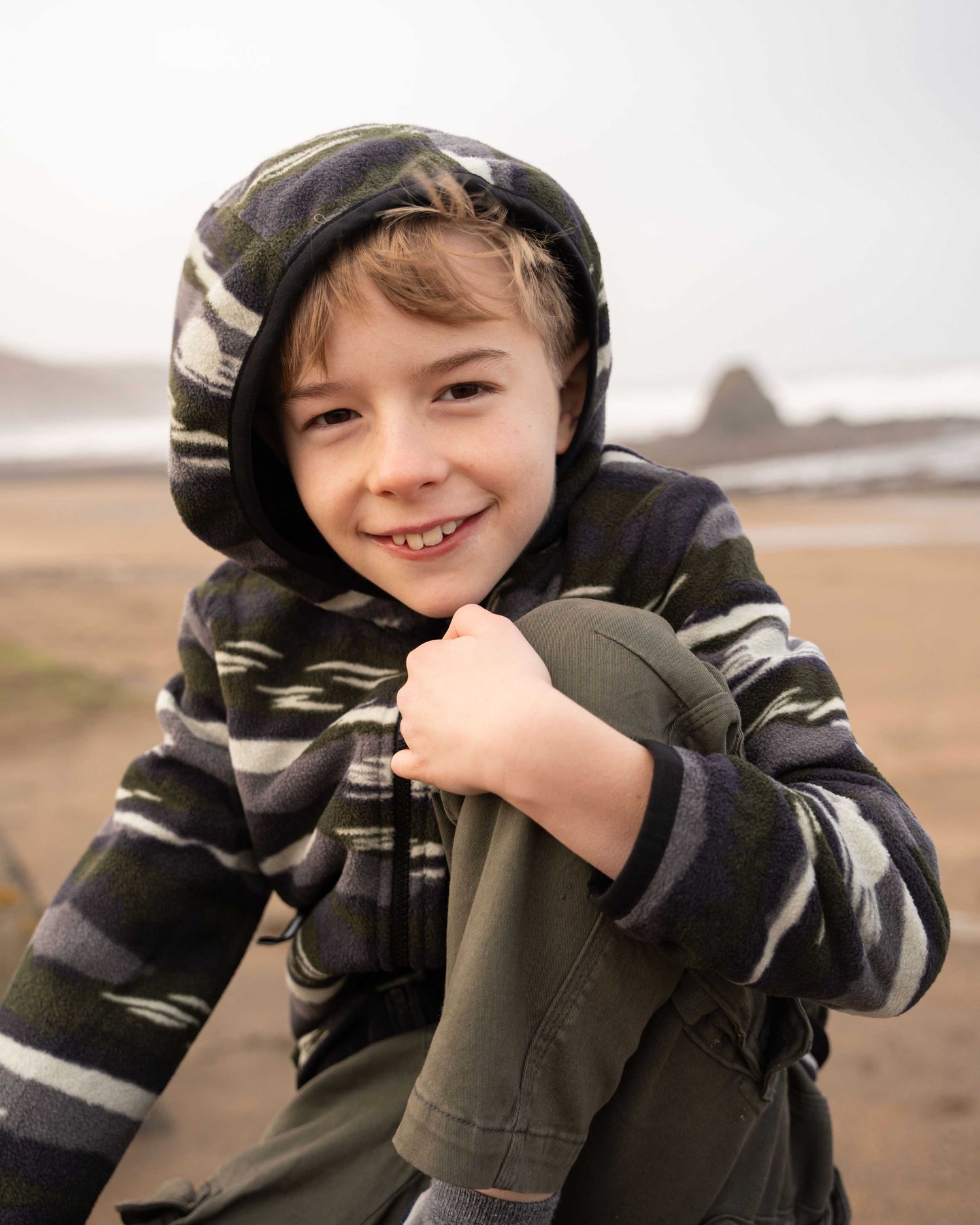 A child wearing the Saltrock Camo Stripe Kids Recycled Micro Fleece in Green sits on a sandy beach, smiling with a rocky coastline in the background.