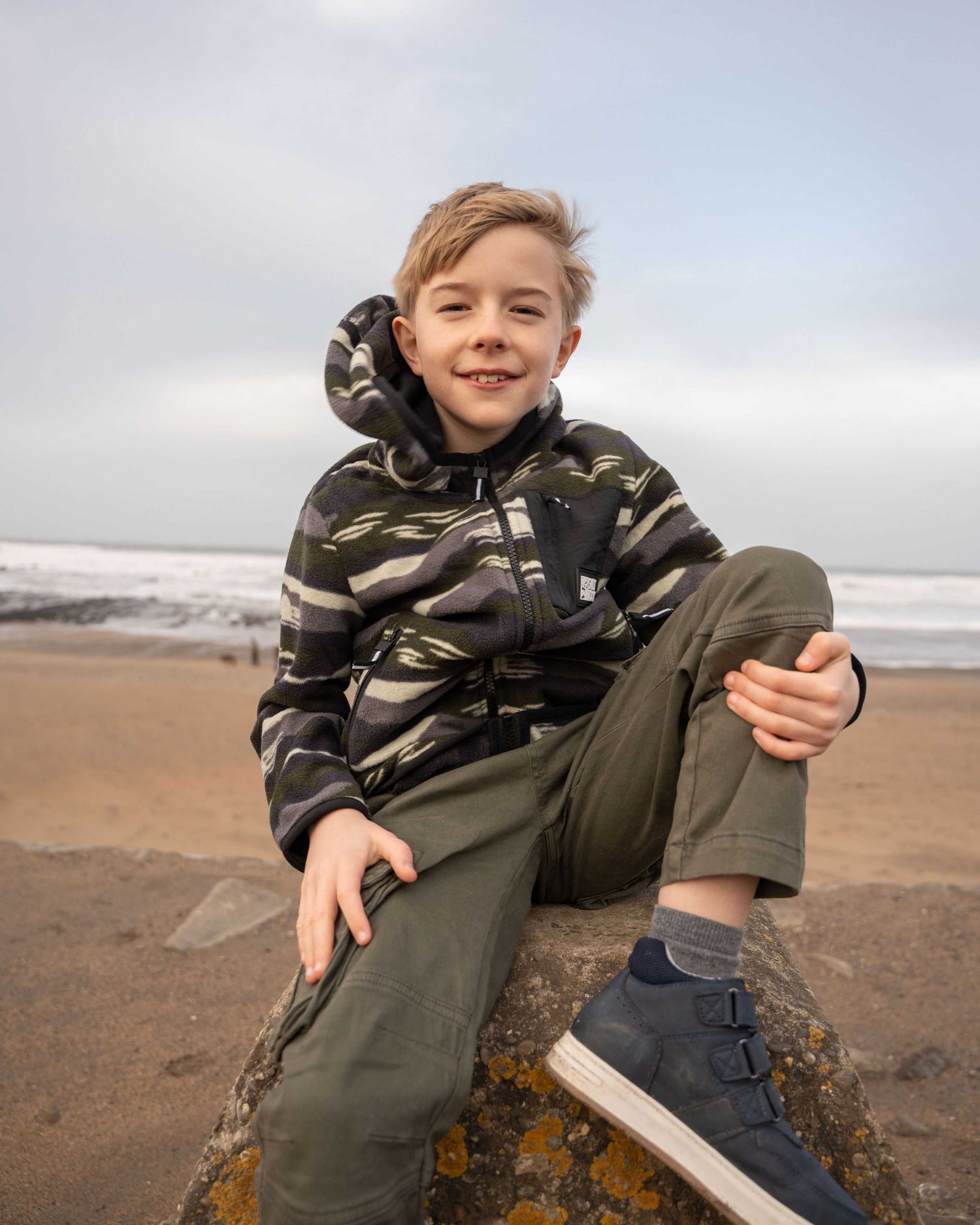 A child wearing a Saltrock Camo Stripe Kids Recycled Micro Fleece in green sits on a rock by the beach, smiling against a backdrop of ocean waves and a cloudy sky.