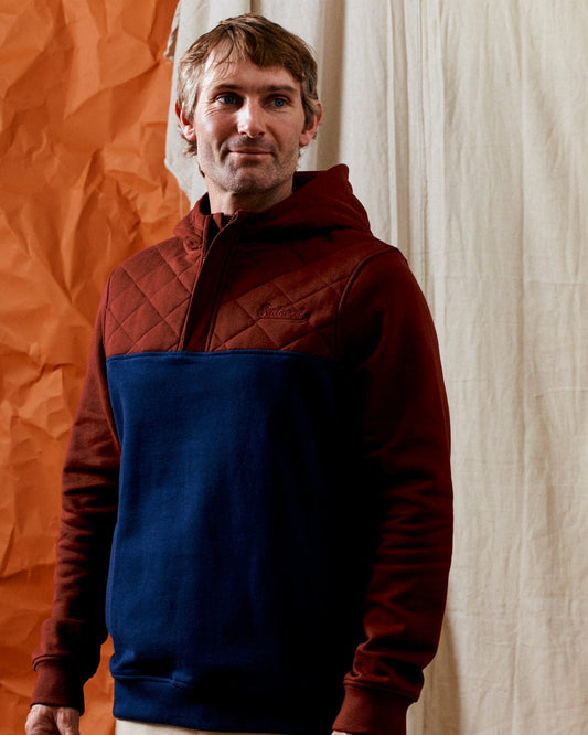 A man wearing a Saltrock Aiken men's 1/4 neck recycled hoodie with a red quilted top and navy bottom stands against an orange and beige backdrop.