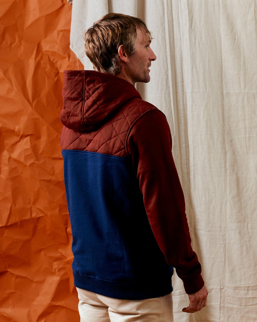 A person wearing the Aiken - Mens 1/4 Neck Recycled Hoodie in red by Saltrock stands with their back facing the camera, against a backdrop of orange and beige textured fabrics.