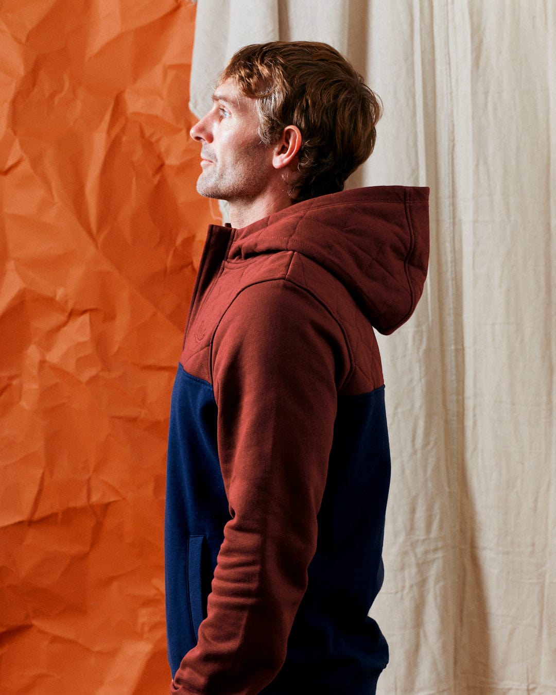 A man wearing the Saltrock Aiken men's 1/4 neck recycled hoodie in red stands in profile against a backdrop of orange and beige crumpled fabric.