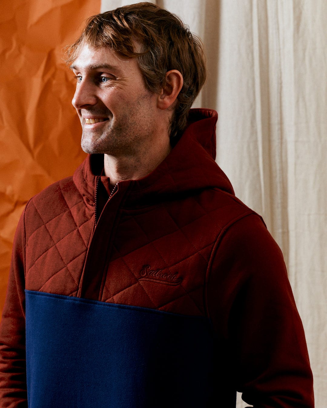 A man in a Saltrock Aiken men's 1/4 neck recycled hoodie in red with a blue section stands smiling in front of a crinkled orange and white backdrop, proudly showcasing the Saltrock branding.