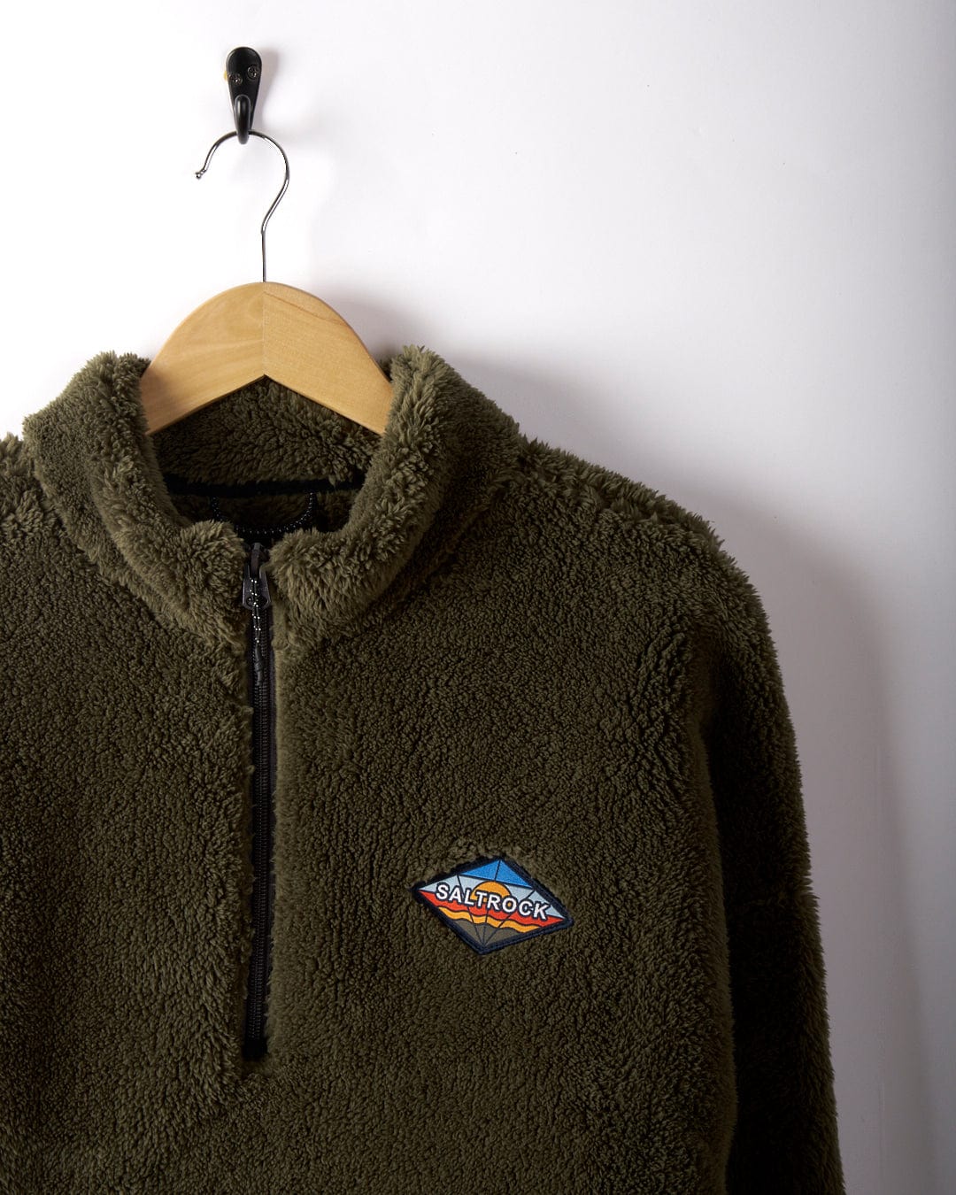 A green Arches men's recycled oversized fleece from Saltrock, featuring a quarter-zip and an embroidered logo, hangs on a wooden hanger against a white wall.