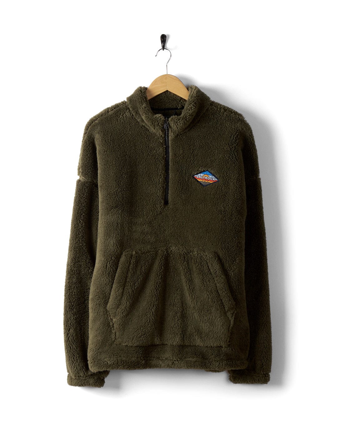 Hanging on a wooden hanger against a white background is the Arches - Men's Recycled Oversized Fleece in Green by Saltrock, featuring an oversized fit, half-zip front, kangaroo pocket, and a small colorful patch on the left chest.