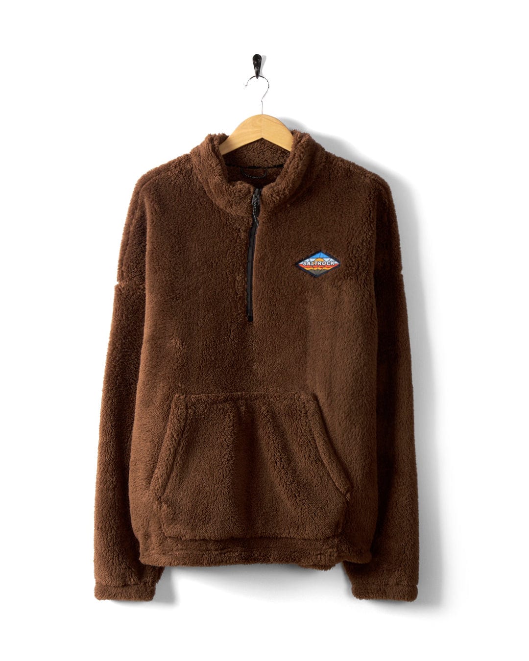 The Arches Men's Recycled Oversized Fleece by Saltrock is a brown, fuzzy zip-up sweatshirt made from super soft teddy fleece. It features a front pocket and a small embroidered patch on the left chest, and is displayed hanging on a wooden hanger against a white background.