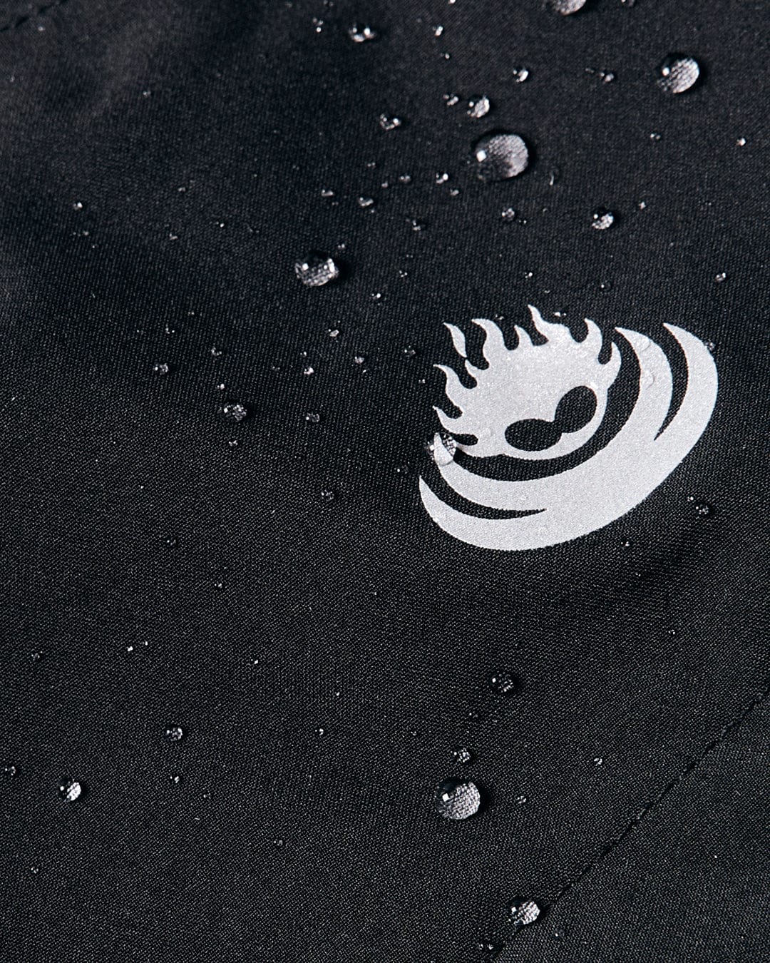 Close-up of water droplets on the black, waterproof polyester fabric of the Whistler Men's Hooded Jacket by Saltrock, showcasing a white abstract logo design with a flame and heart shape.