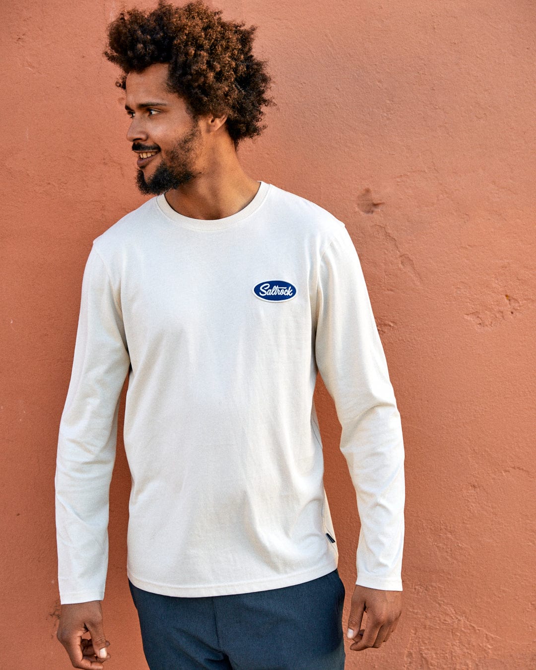 A man with curly hair smiling and leaning against an orange wall, wearing a white long-sleeve shirt with a blue Saltrock badge.