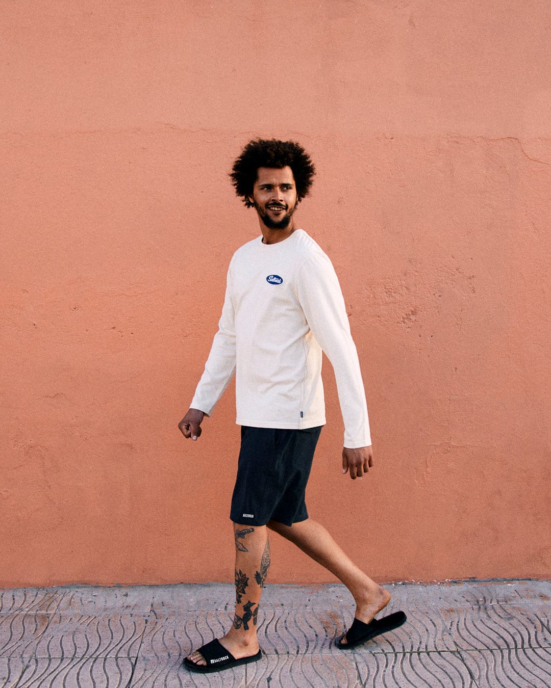 A young man with curly hair, wearing a Saltrock cream long-sleeve t-shirt and crew neckline, shorts, and sandals, walking past a pink wall.