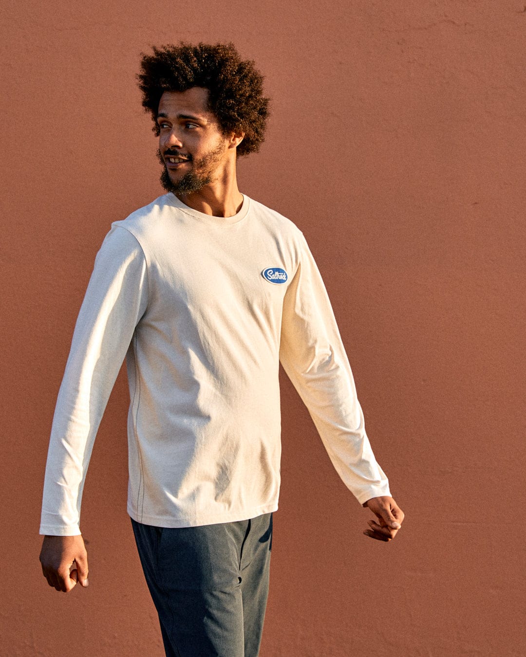 A young man with an afro wearing a Saltrock Attendant Mens Long Sleeve T-Shirt in Cream, standing against a salmon-colored wall, looking to the side with a relaxed posture.