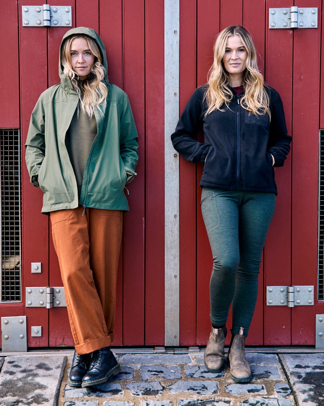 Two people with long hair stand against a red, metal door. One is wearing a green jacket made from recycled yarns and orange pants, while the other is sporting Saltrock's Ava women's fleece in black and green pants, both in standard fit.