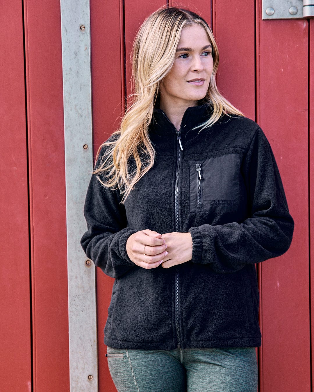 A person wearing the Ava Women's Fleece in black from Saltrock, crafted with recycled yarns, stands in front of a red wall.