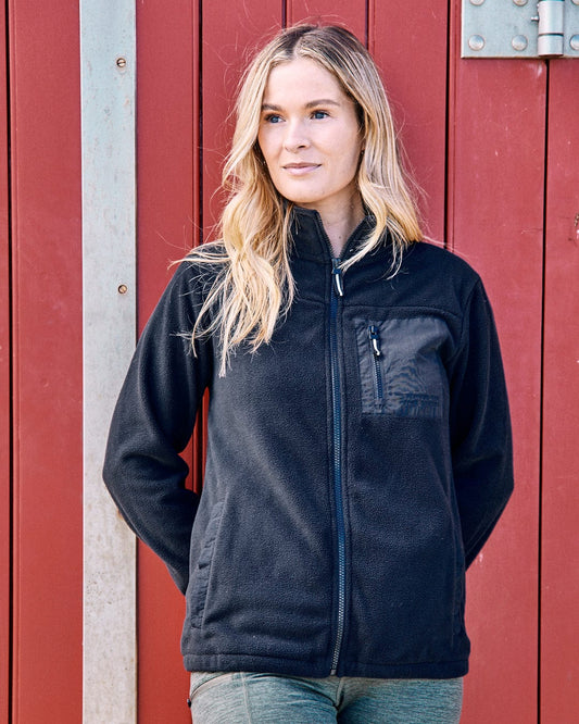 A person stands in front of a red wooden wall, wearing the Saltrock "Ava" women's fleece in black, hands behind their back.