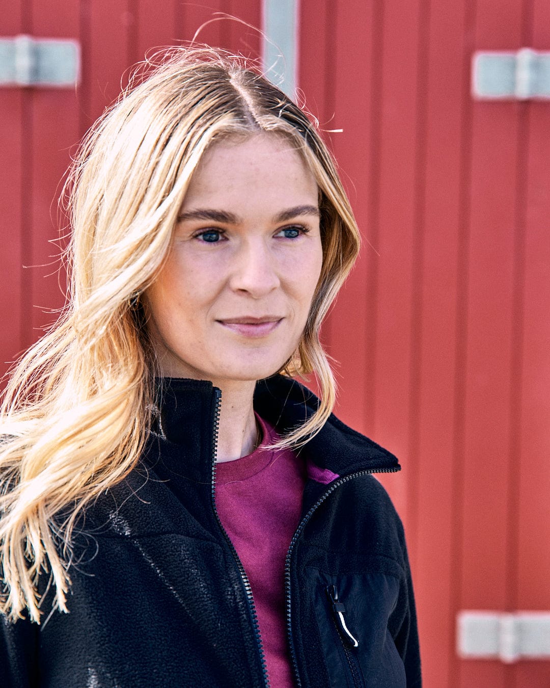 A person with long blond hair wearing the Ava women's fleece from Saltrock in black and a maroon top stands in front of a red wooden background.