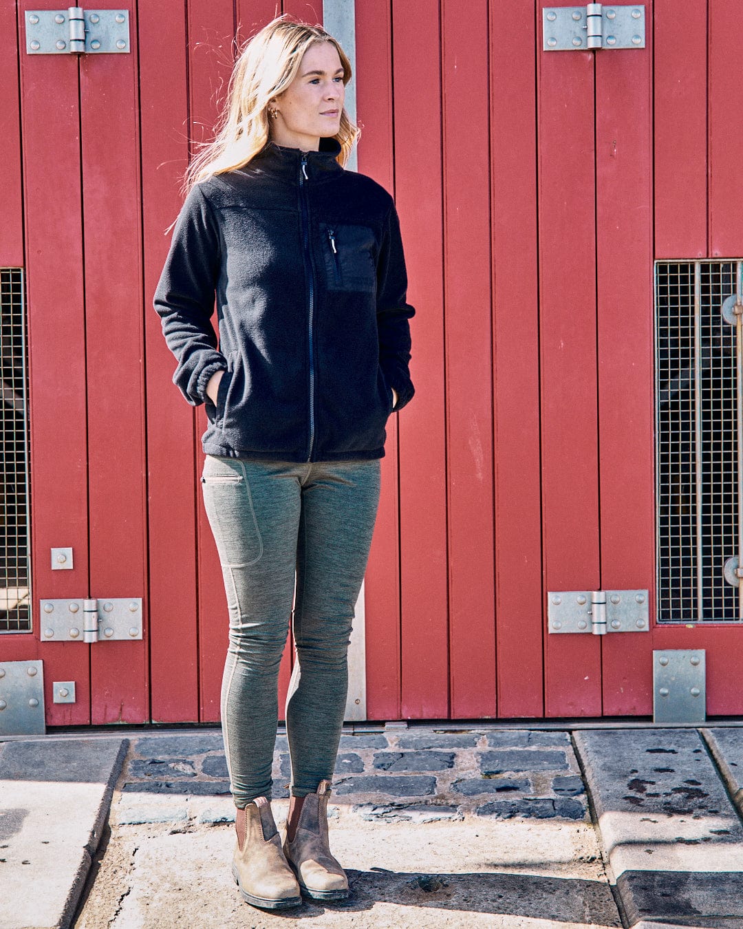 Standing in front of a red door, the person showcases the Ava Women's Fleece in black by Saltrock, paired with green pants and brown boots.