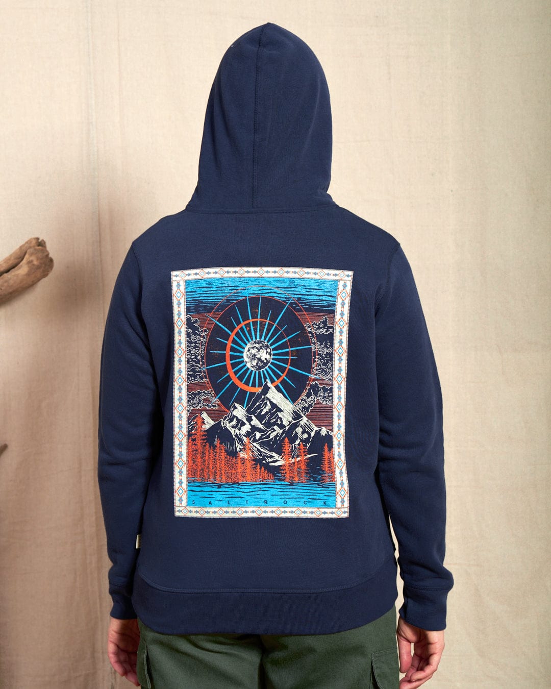 A person wearing the Saltrock Aztec Mountain Women's Zip Hoodie in blue features a vibrant design with mountains, trees, and a sunburst. The cosmic mountain scene on the back complements the drawstring hood, while subtle Saltrock branding adds stylish flair.