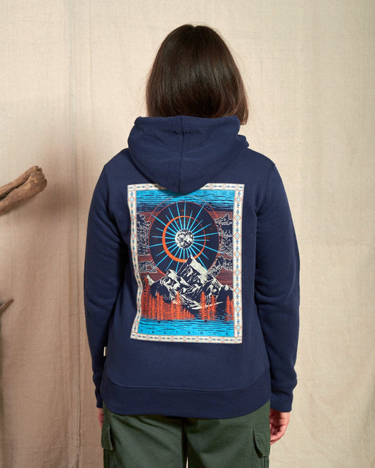 A person indoors wears the Saltrock Aztec Mountain Women's Zip Hoodie in blue, showcasing a cosmic mountain scene with a sunburst design on the back. The hood with drawstrings enhances comfort, while subtle Saltrock branding completes the look.