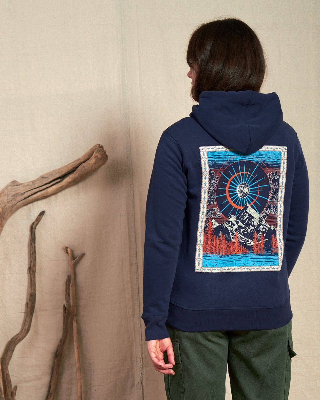 A woman in a Saltrock Aztec Mountain blue zip hoodie, featuring a colorful cosmic mountain scene on the back and navy drawstrings, stands against a canvas backdrop with driftwood leaning nearby.