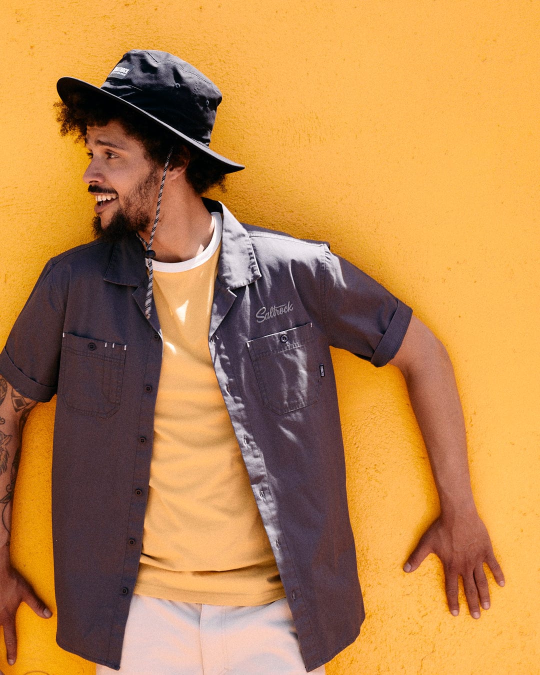 A man in a Saltrock Boulevard - Mens Short Utility Sleeve Shirt - Dark Grey and a dark grey bucket hat leans against a yellow wall, looking to the side and smiling, exuding utility worker style.