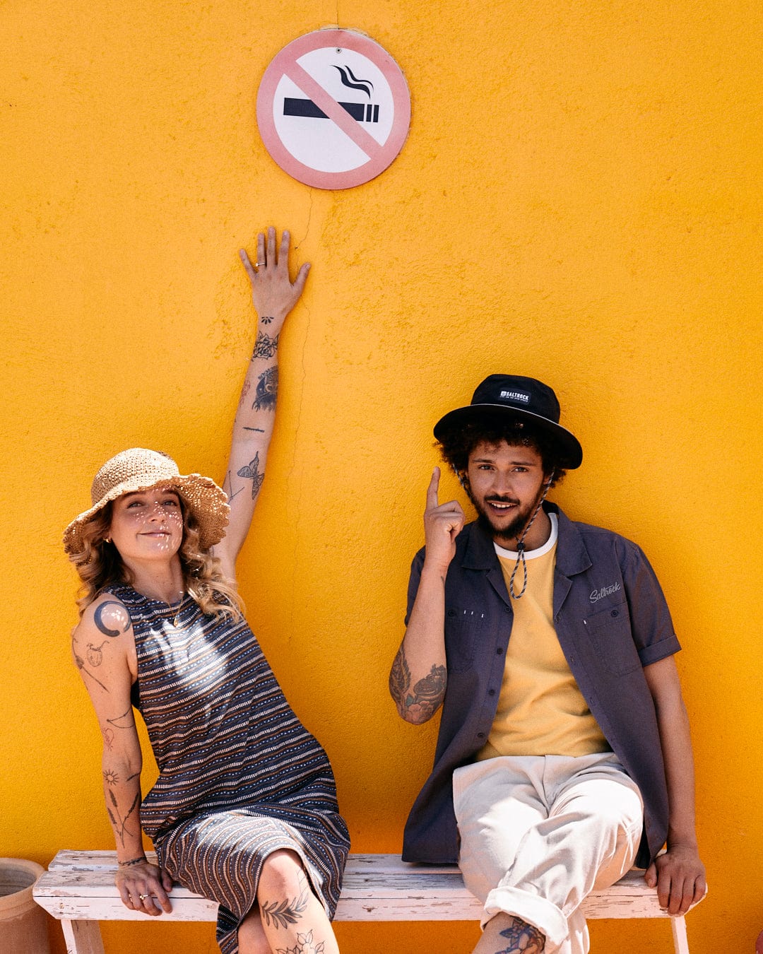 A woman in a striped dress and sun hat points to a no-smoking sign, while a man in Saltrock's Boulevard - Mens Short Utility Sleeve Shirt - Dark Grey sits next to her, gesturing and smiling, in front of a bright yellow wall.