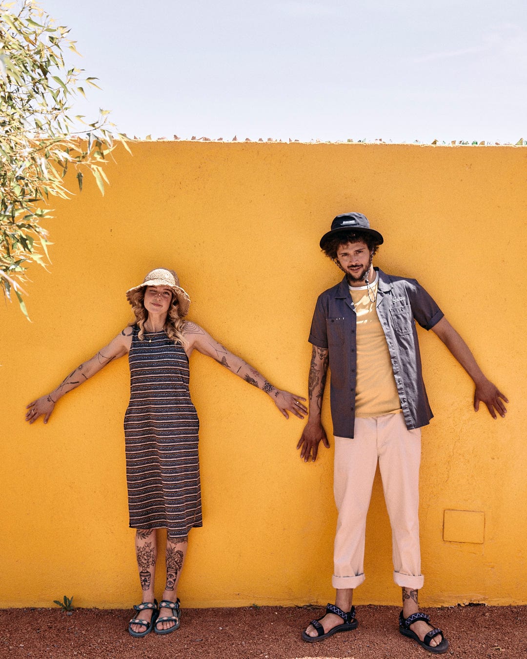 Two people stand with arms spread against a bright yellow wall. One wears a striped dress and sunhat, the other sports utility worker style, featuring a hat, gray shirt (Saltrock Boulevard - Mens Short Utility Sleeve Shirt - Dark Grey), and beige pants. Both have tattoos and are wearing sandals.