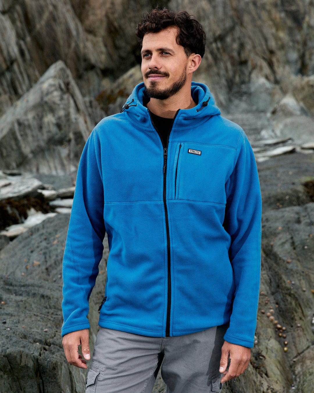 A person wearing the Saltrock Bradford Men's Recycled Hooded Fleece in blue stands against a rocky outdoor background.