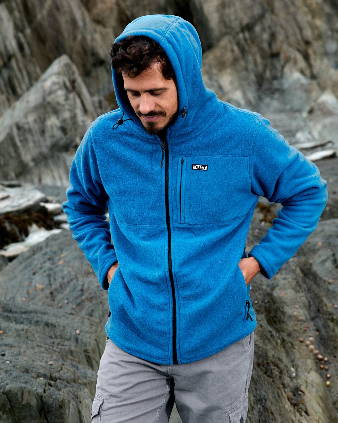 A man wearing the Saltrock Bradford men's recycled blue hooded fleece stands on a rocky outdoor surface.