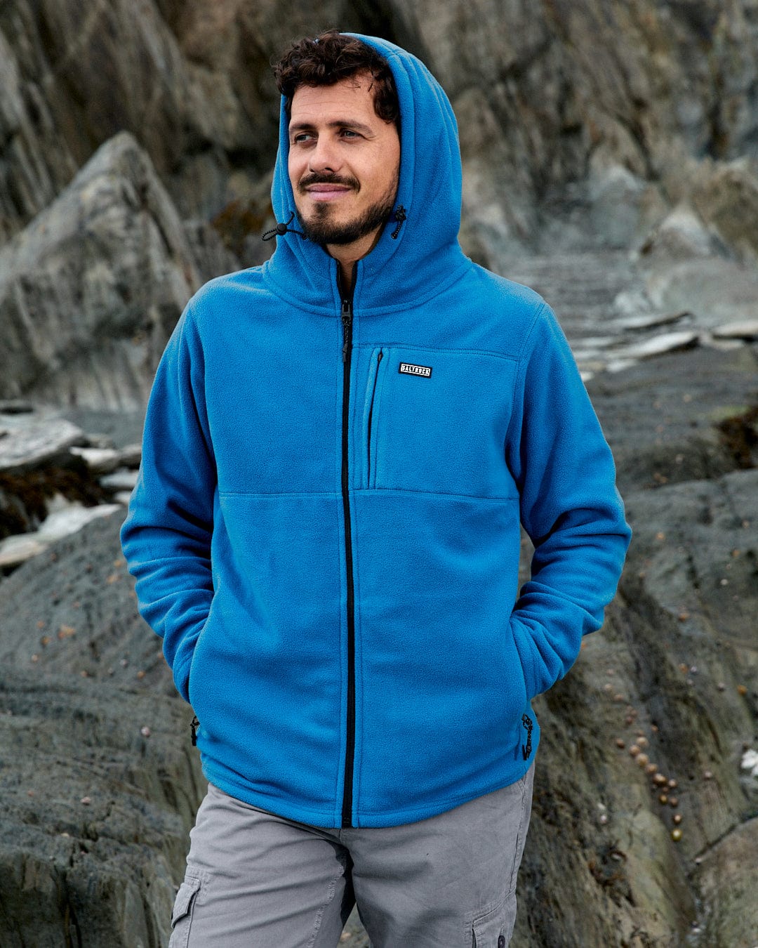 A person stands outdoors wearing the Saltrock Bradford recycled hooded fleece in blue, set against a backdrop of rocky terrain.