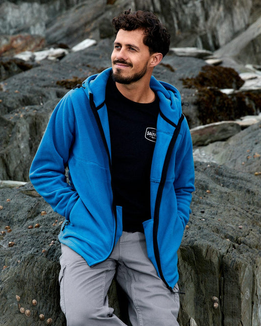 A man in the Saltrock Bradford Men's Recycled Hooded Fleece in blue and gray pants stands on rocky terrain, gazing to the side.