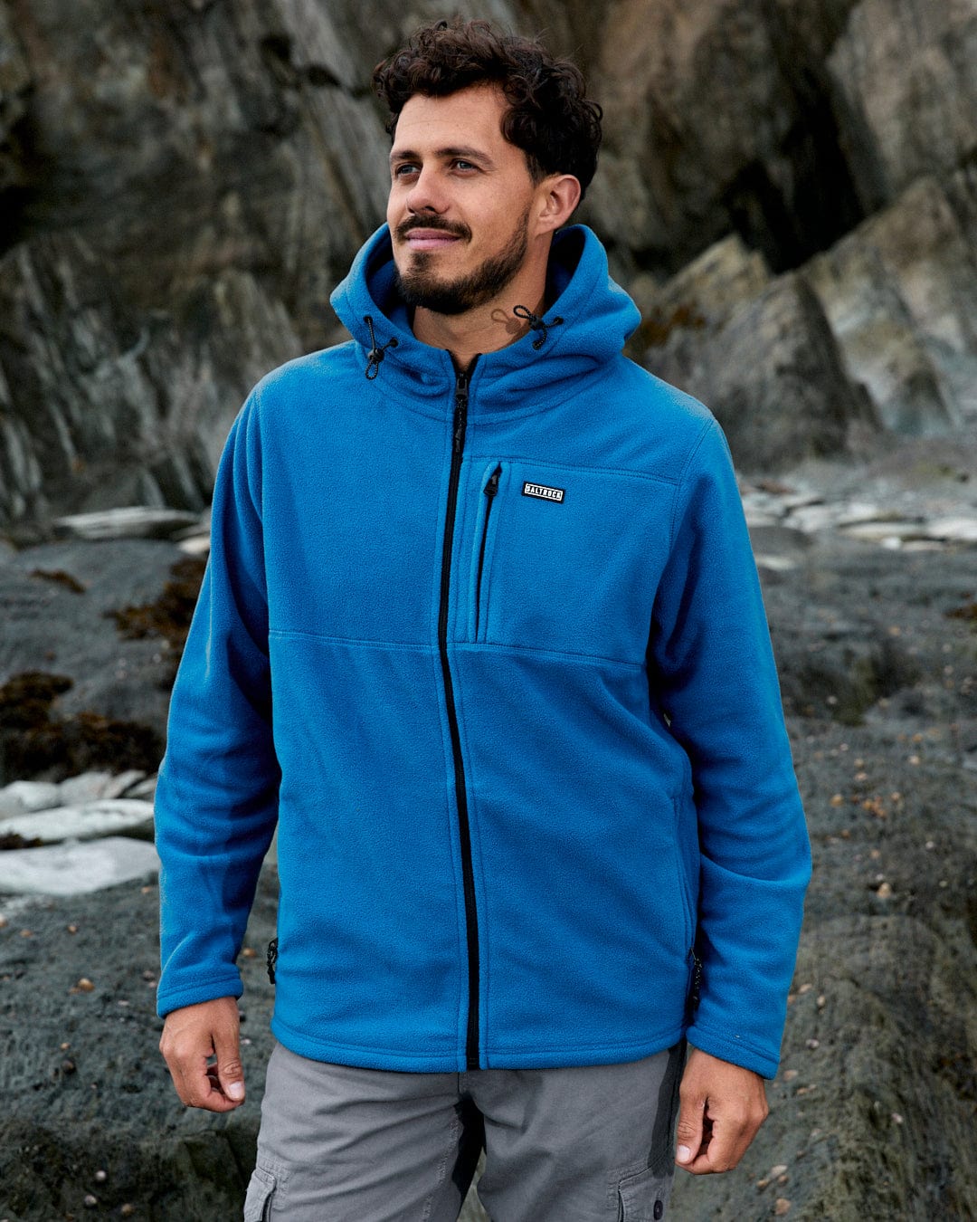 A person wearing the Saltrock Bradford Men's Recycled Hooded Fleece in blue stands outdoors in front of rocky terrain.