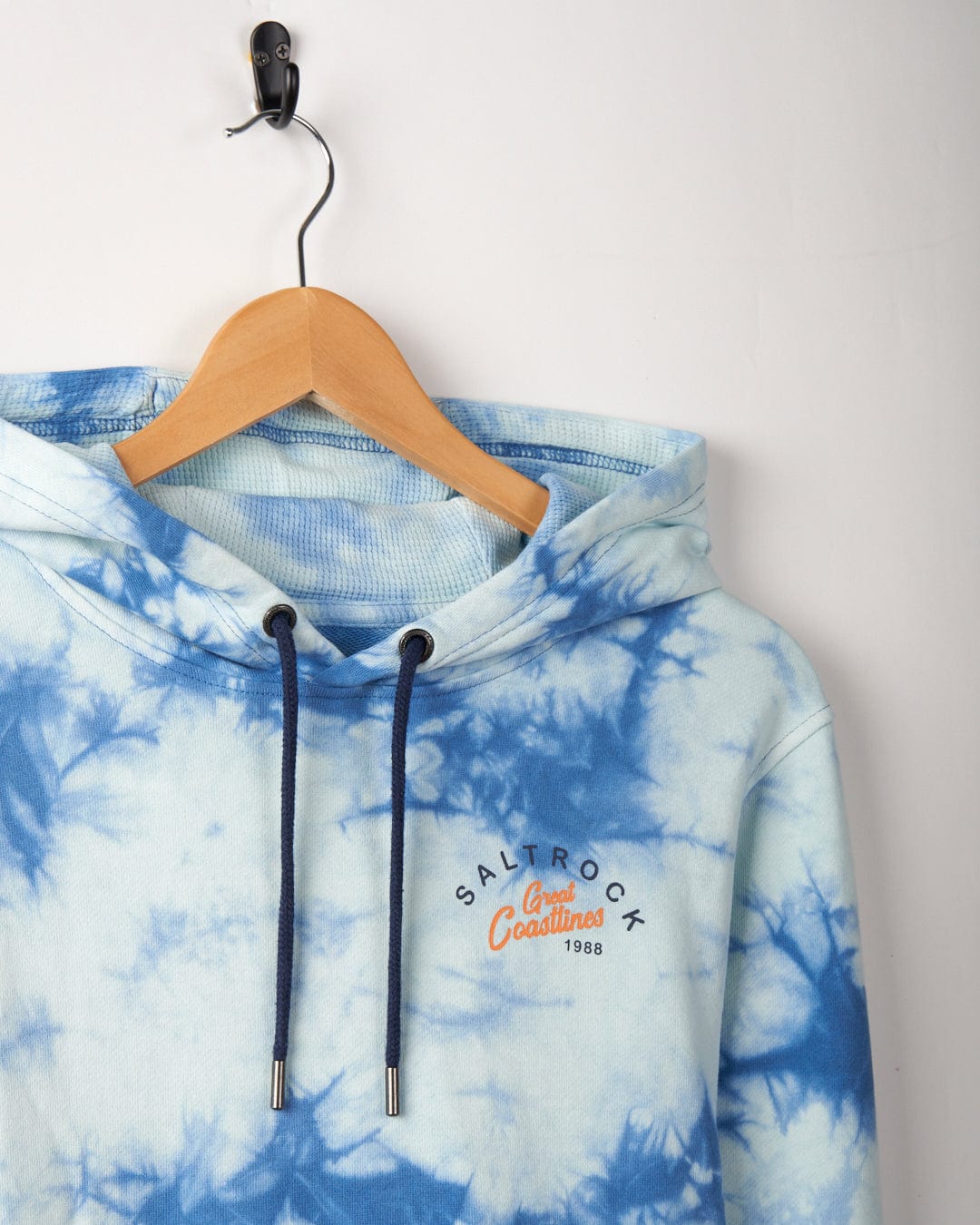 A Coastline - Mens Tie Dye Pop Hoodie - Blue made from 100% cotton with the logo "Saltrock Life" and text "1988 Coastlines," hanging on a wooden hanger against a white wall.