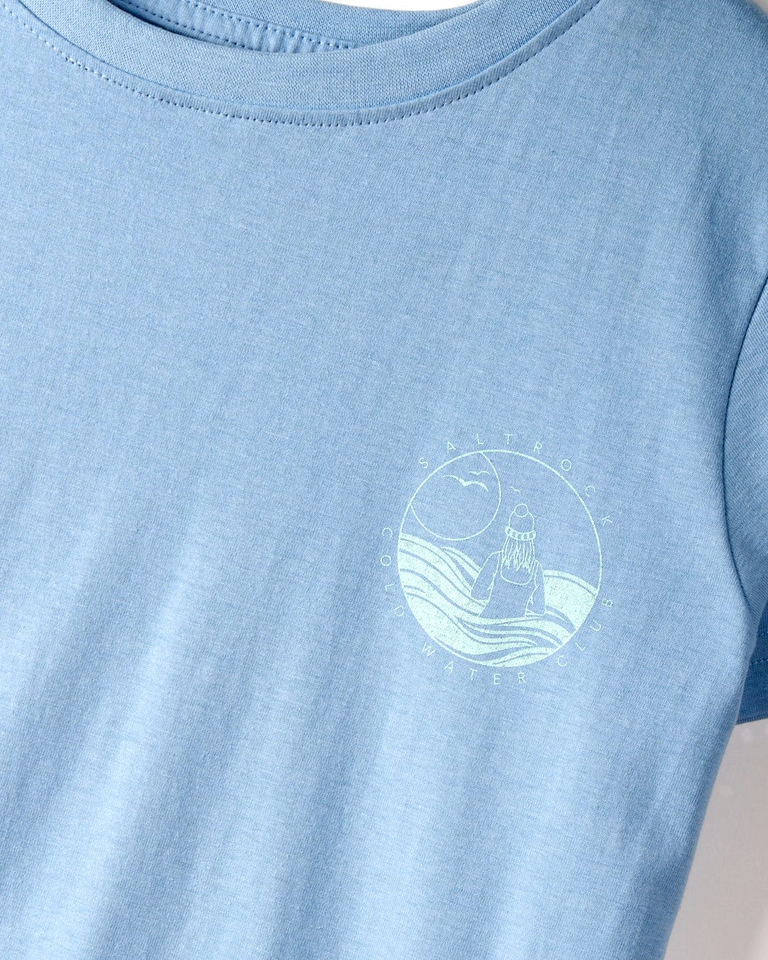 Close-up of a light blue t-shirt made from 100% cotton, featuring a small circular design on the chest. The design depicts a person sitting by the water with moon and wave elements, labeled "Cold Water Club - Womens T-Shirt - Blue" by Saltrock.