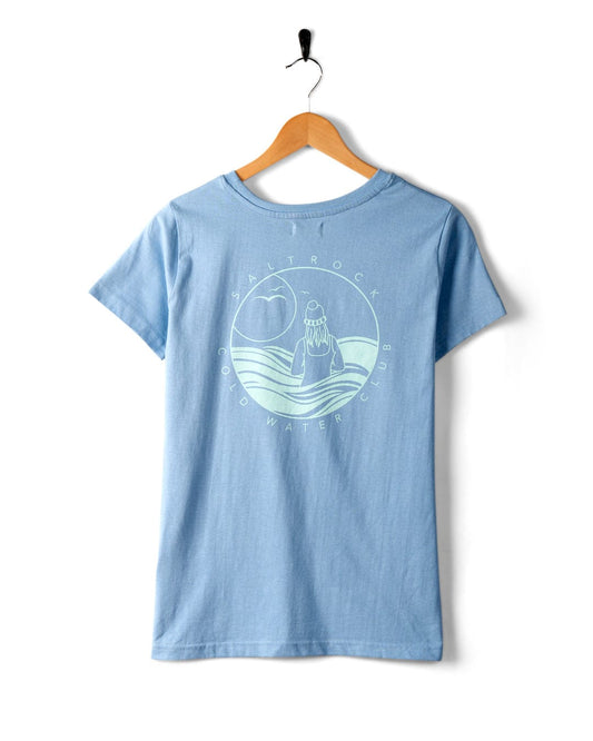 A Cold Water Club - Womens T-Shirt - Blue by Saltrock hangs on a wooden hanger, featuring a circular logo with a person, waves, and the sun. The text reads "Saltrock Cold Water Club.