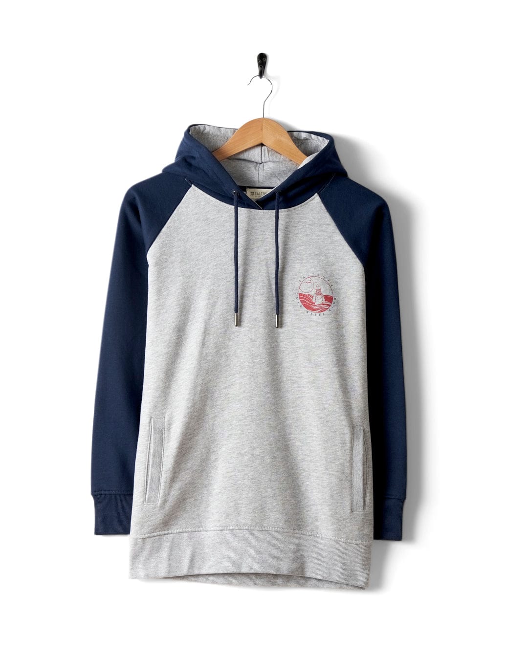 The Saltrock Coldwater Club Women's Raglan Pop Hoodie in grey features navy contrasting sleeves, a small red chest emblem, and hangs on a wooden hanger against a white background.