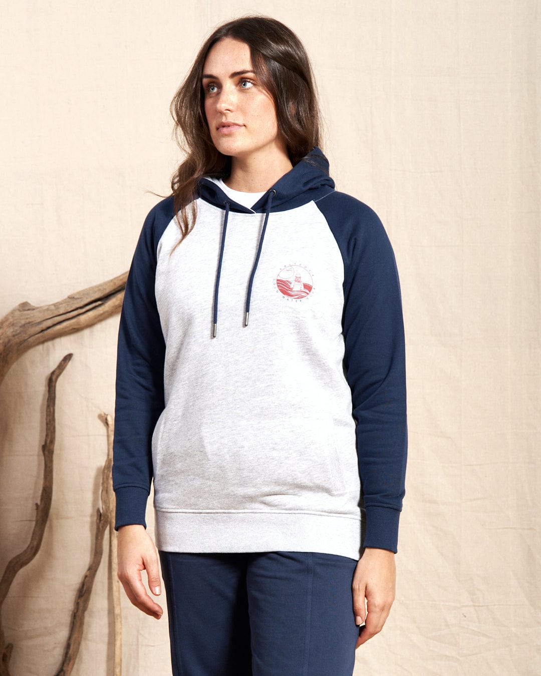 A person in a Saltrock Coldwater Club Women's Raglan Pop Hoodie, featuring gray color with navy accents, stands against a neutral backdrop complemented by driftwood.