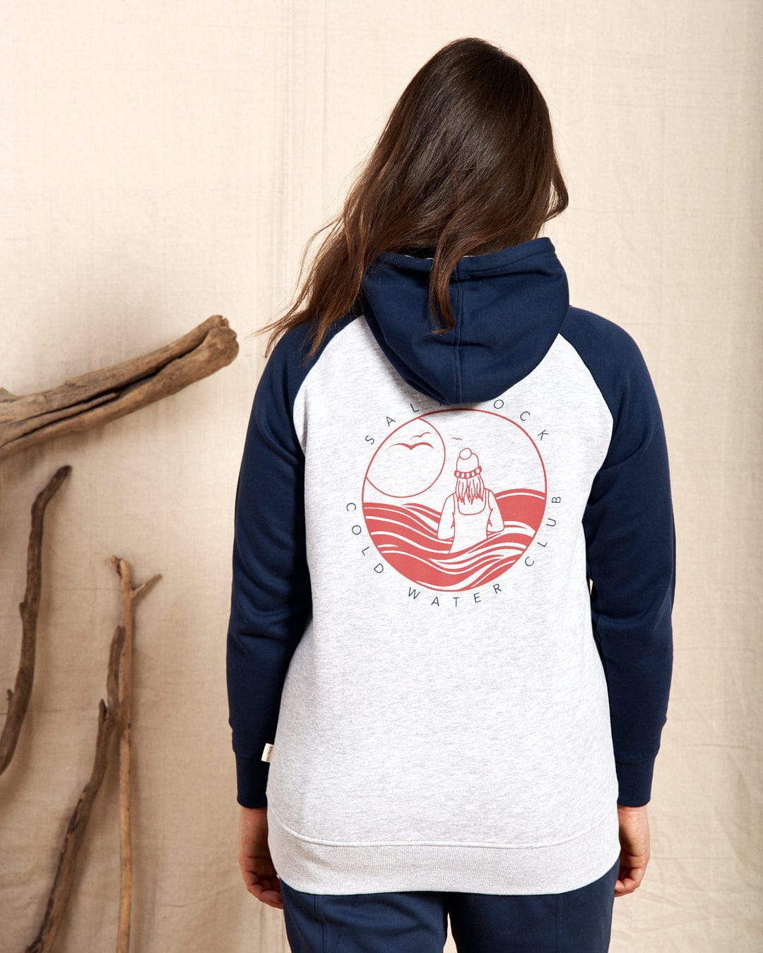 A person wears the Saltrock "Coldwater Club - Womens Raglan Pop Hoodie - Grey," featuring contrasting sleeves and a back design of a figure by water, a rising/setting sun, and "Salt Rock Cold Water Club.